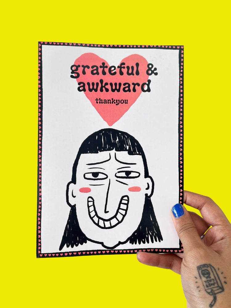 Card with a face illustration and text 'grateful & awkward' held against a yellow background